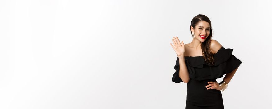 Gorgeous young woman in black dress, earrings and jewelry, waving hand and smiling friendly, saying hello, standing over white background.