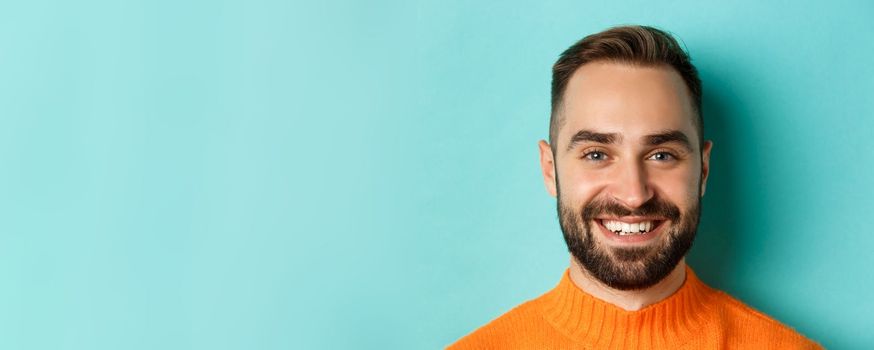 Headshot of handsome caucasian man with beard smiling happy at camera, standing in orange sweater against turquoise background.