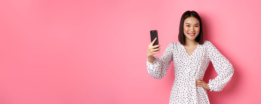 Beautiful asian girl using photo filters app and taking selfie on mobile phone, posing in cute dress against pink background.