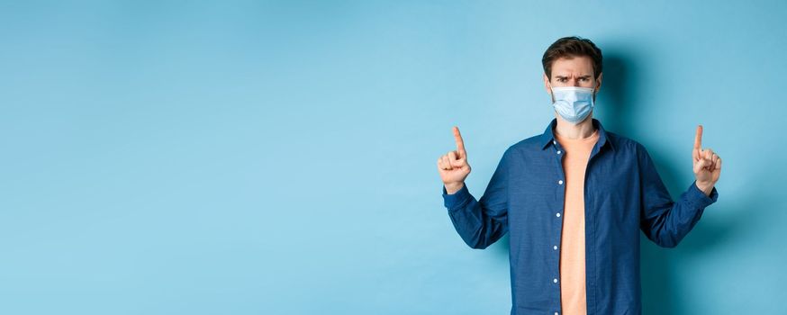 Covid and healthcare concept. Angry man in face mask pointing fingers up, feel disappointed and upset, standing on blue background.