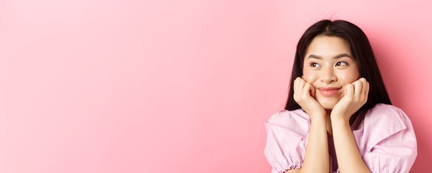 Close-up of excited asian girl listening with interest, smiling amused and looking aside at logo, daydreaming, standing against pink background.