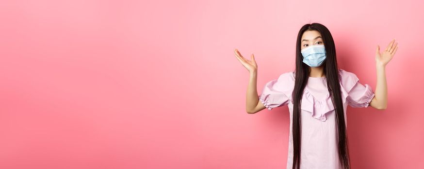 Coronavirus, quarantine and lifestyle concept. Clueless asian girl in face mask shrugging shoulders unware, know nothing, standing on pink background.