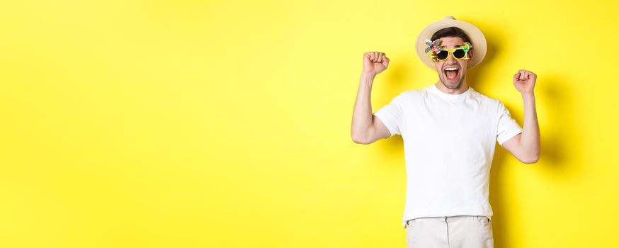 Concept of tourism and lifestyle. Happy man winning trip to resort, shouting yes and raising hands up, triumphing, wearing sunglasses and summer hat, yellow background.