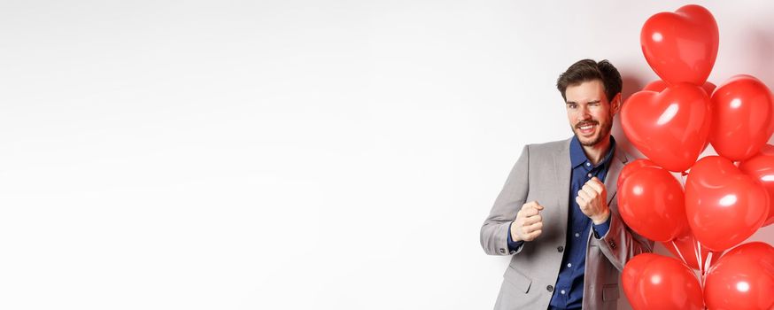 Handsome man dancing and smiling near Valentines day hearts balloon, winking at camera, getting dressed on romantic date on lovers holiday, white background.