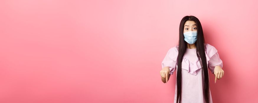 Healthy people and covid-19 pandemic concept. Excited asian girl in face mask pointing fingers down, showing advertisement with interest, standing against pink background.