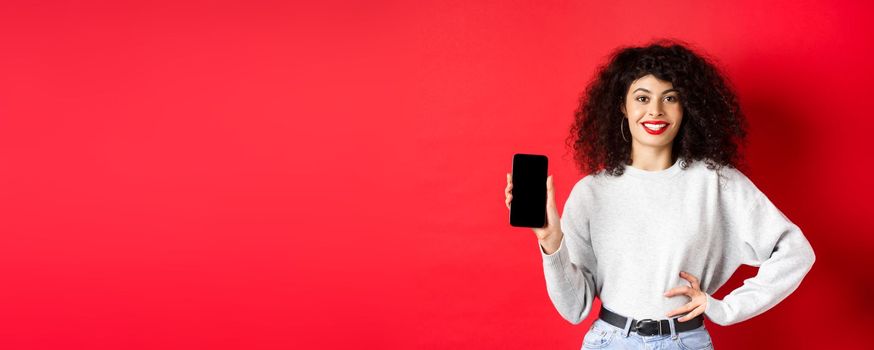 Attractive smiling woman showing empty smartphone screen and looking happy, advertising online store or application, standing against red background.