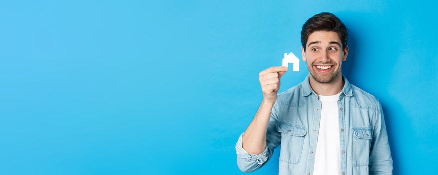Real estate concept. Excited guy looking at small house model and smiling, renting apartment, standing over blue background.