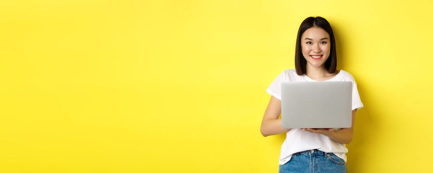 Cute asian woman studying on laptop and smiling, standing in white t-shirt and jeans against yellow background.