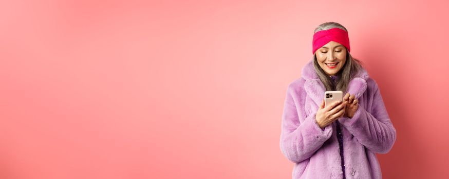 Online shopping and fashion concept. Modern asian granny in trendy faux fur coat texting message, using smartphone and looking happy at screen, pink background.