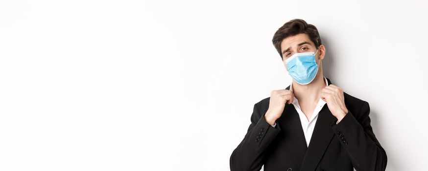 Concept of covid-19, business and social distancing. Image of confident handsome man in trendy suit and medical mask, looking sassy, standing against white background.