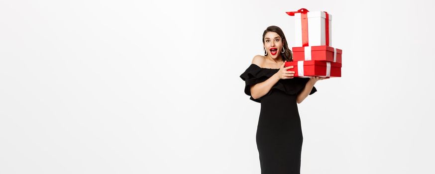 Merry christmas and new year holidays concept. Cheerful lady in black dress holding xmas presents and smiling at camera, standing over white background.