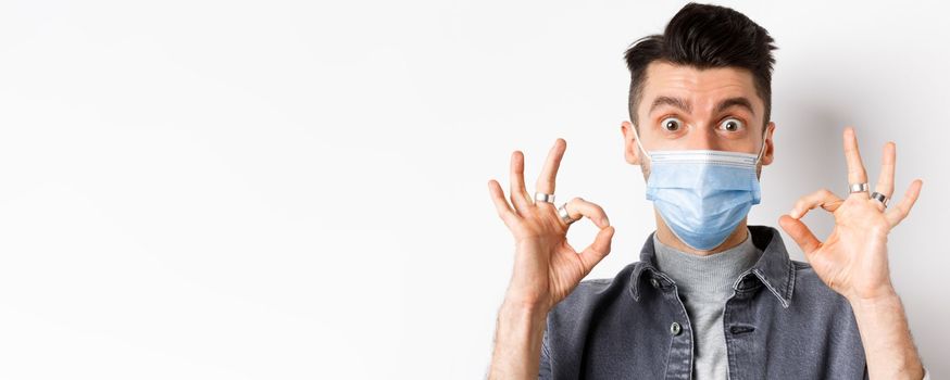 Covid-19, health and lifestyle concept. Close-up of excited man in face mask show okay signs and look amazed, praise good thing, standing on white background.