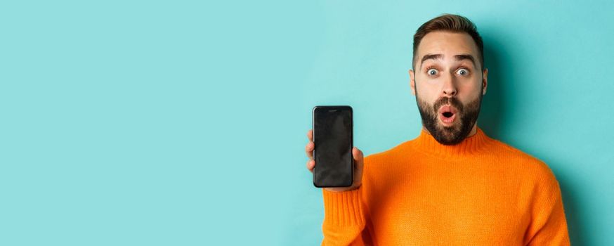 Close-up of handsome bearded guy in orange sweater, showing smartphone screen and smiling, showing promo online, turquoise background.