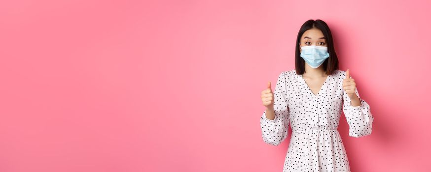 Covid-19, quarantine and lifestyle concept. Excited asian woman in face mask showing thumbs-up, praising good job, looking amazed at camera, standing over pink background.