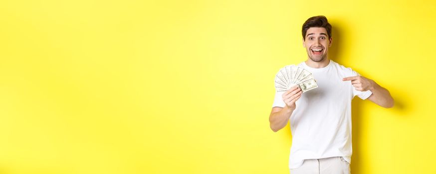 Happy handsome guy pointing finger at money, concept of credit and loan, standing over yellow background.