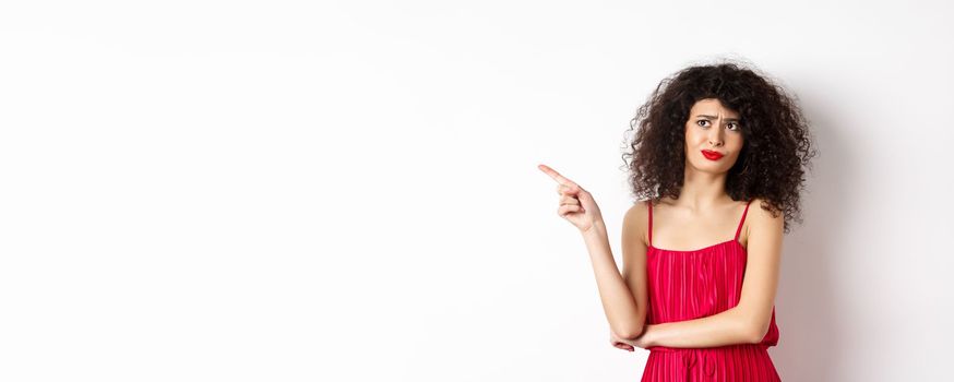 Disappointed and skeptical young woman with curly hair, wearing red dress, grimacing and pointing finger left at logo, standing over white background.