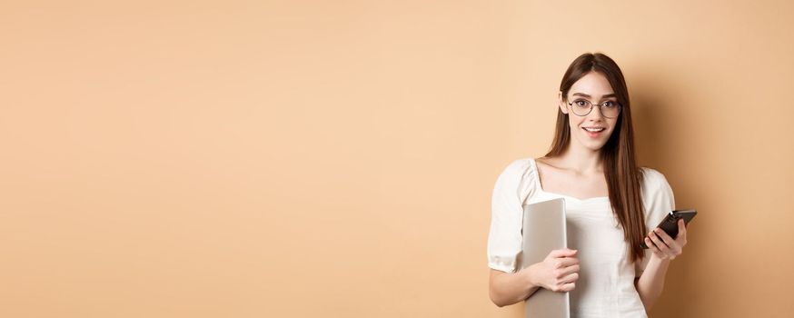 Smiling young woman in glasses reading exciting news on mobile phone, holding laptop and looking happy at camera, standing on beige background.
