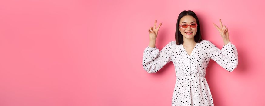 Tourism and lifestyle concept. Beautiful asian woman on summer vacation, showing peace signs and smiling happy, wearing trendy sunglasses, pink background.
