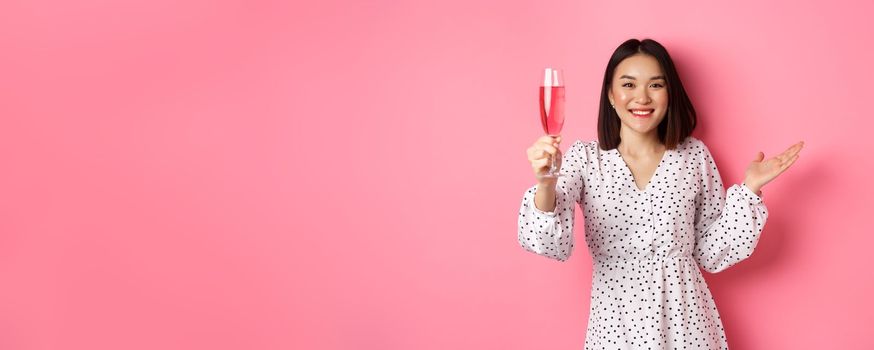 Beautiful asian woman raising glass of champagne and looking happy at camera, congratulating you on party, celebrating, standing over pink background.