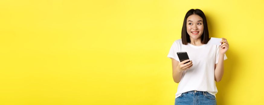 E-commerce and online shopping concept. Cheerful asian girl paying in internet, holding smartphone and plastic credit card, smiling and looking left, yellow background.