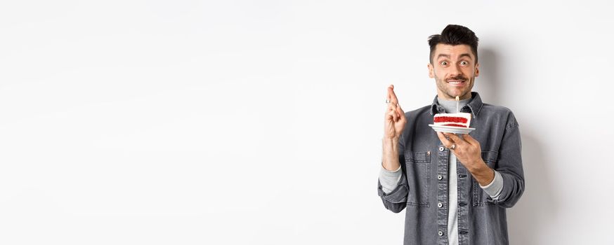 Birthday. Happy guy making wish with fingers crossed, holding bday cake with candle, standing on white background.