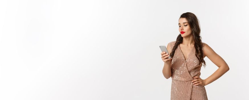 Christmas party and celebration concept. Careless woman in stylish dress reading message on mobile phone, looking skeptical, standing over white background.