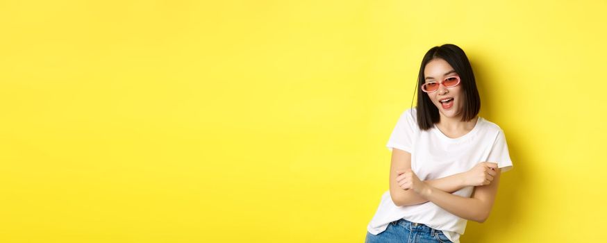 Fashion and lifestyle concept. Sassy and confident asian woman in trendy sunglasses looking self-assured at camera, standing over yellow background.