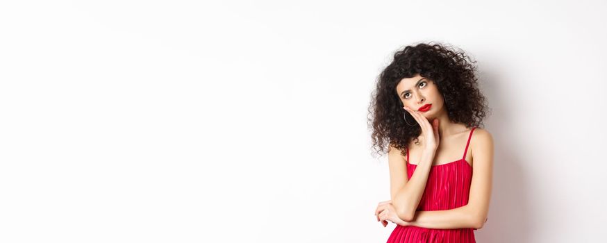 Annoyed and bored young woman with curly hair, look away distressed, lean face on hand, standing bothered in red dress on white background.