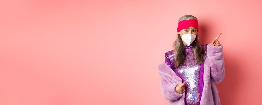 Coronavirus and shopping concept. Worried asian senior woman in stylish outfit and respirator pointing upper right corner, looking unsure at camera, asking advice, pink background.