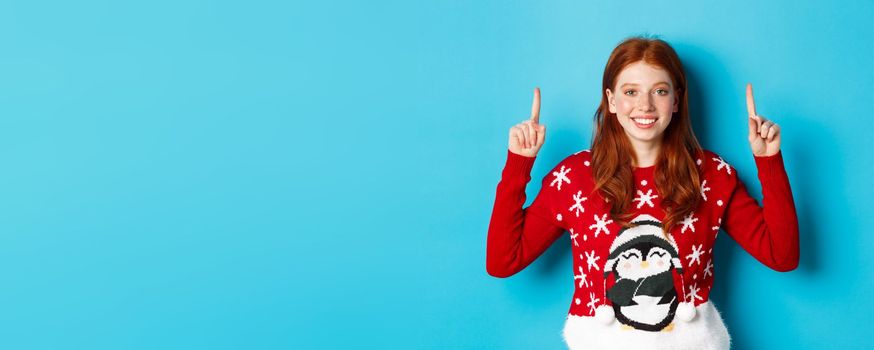 Winter holidays and celebration concept. Cute redhead girl in Christmas sweater, smiling and pointing fingers up at promo logo, standing over blue background.