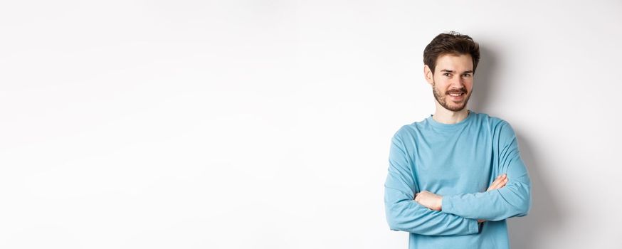 Confident young man with beard, cross arms on chest and smile at camera, standing like professional over white background.