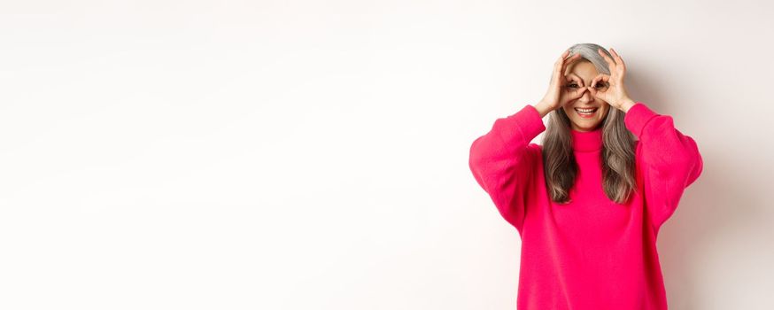 Happy and funny asian senior woman making hand binoculars, looking through fingers and smiling, standing in pink sweater over white background.