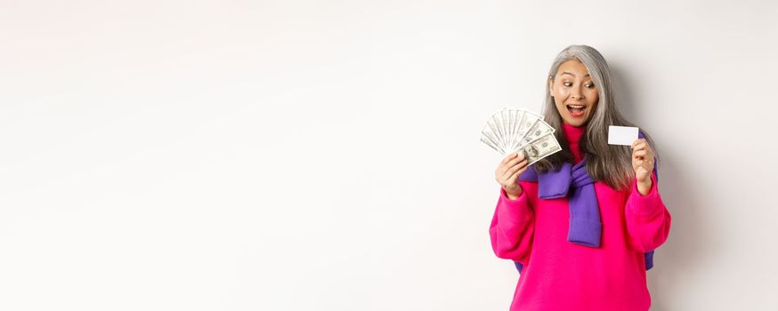 Shopping concept. Lucky asian senior woman looking amazed at credit card and showing money dollars, standing over white background.