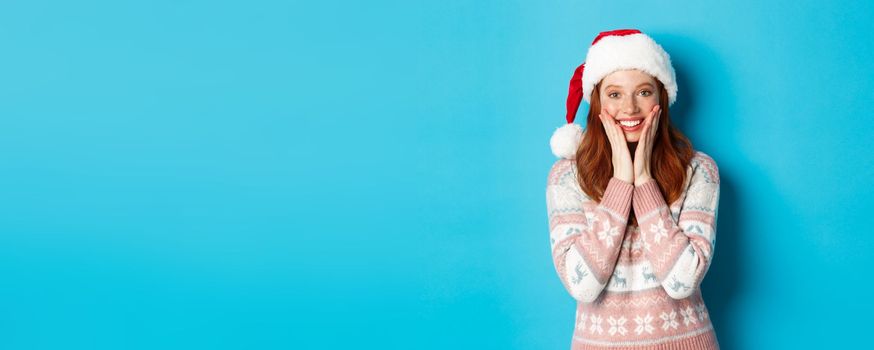 Winter and Christmas Eve concept. Happy redhead girl celebrating xmas, staring at camera amazed, smiling and touching cheeks, standing over blue background.