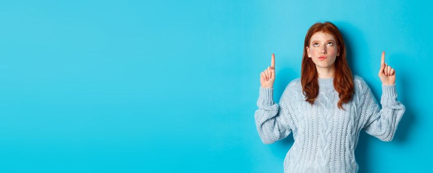 Winter holidays and people concept. Thoughtful redhead girl in sweater staring and pointing fingers up, having doubts, thinking or making choice, standing over blue background.