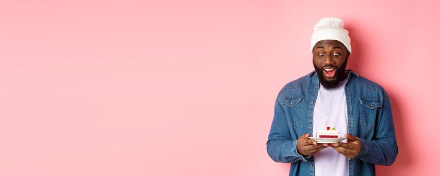 Cheerful african-american guy celebrating birthday, making wish on bday cake with lit candle, smiling happy, standing over pink background.