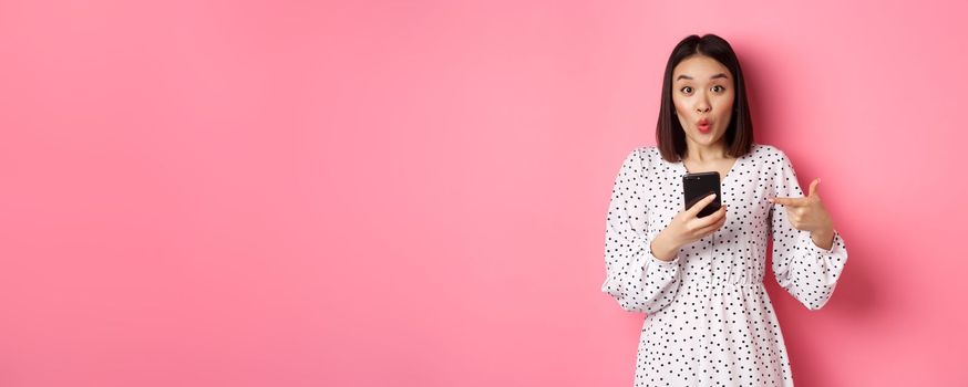 Online shopping and beauty concept. Amazed asian woman pointing finger at mobile phone, recommending smartphone app, standing over pink background.