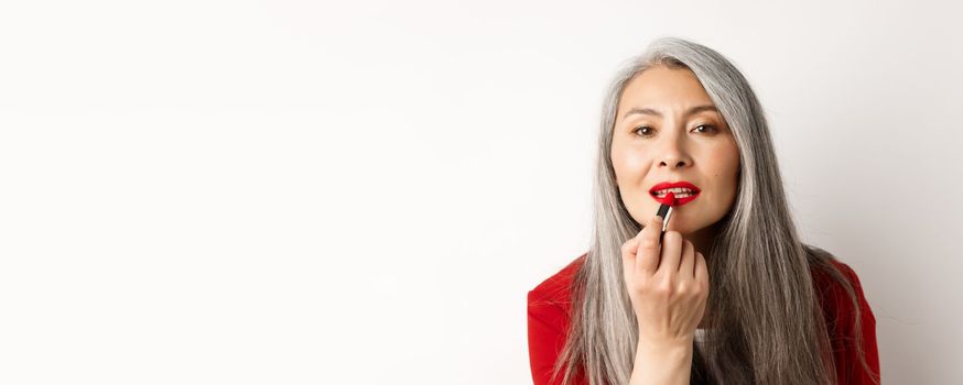 Beauty and fashion concept. Stylish asian mature woman with grey hair, looking in mirror and apply red lipstick, standing over white background.