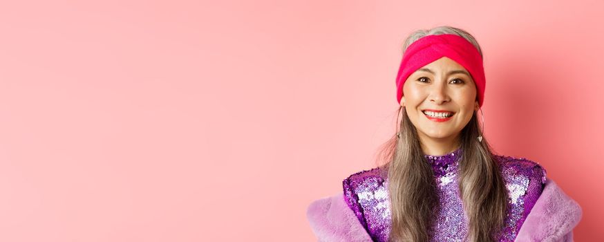 Close-up of trendy asian senior woman with grey hair and stylish disco dress, smiling happy at camera, standing over pink background.