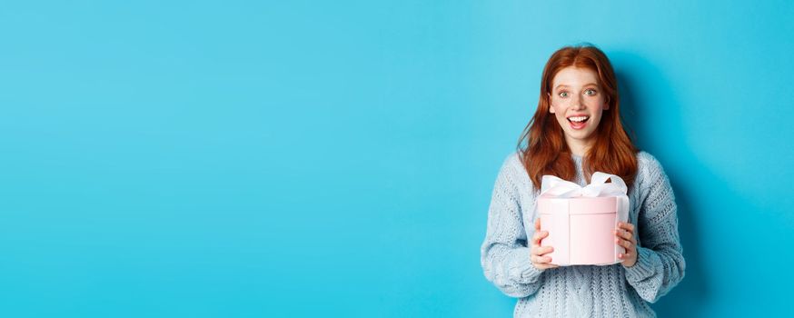 Surprised redhead girl receiving valentines gift, holding box with present and staring at camera amazed, wearing sweater, standing over blue background.