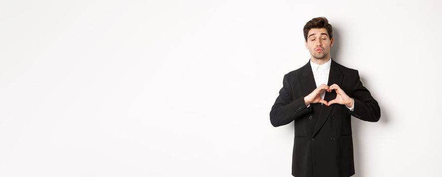 Image of handsome groom in black suit, showing heart sign, close eyes and pucker lips, waiting for kiss, standing against white background.