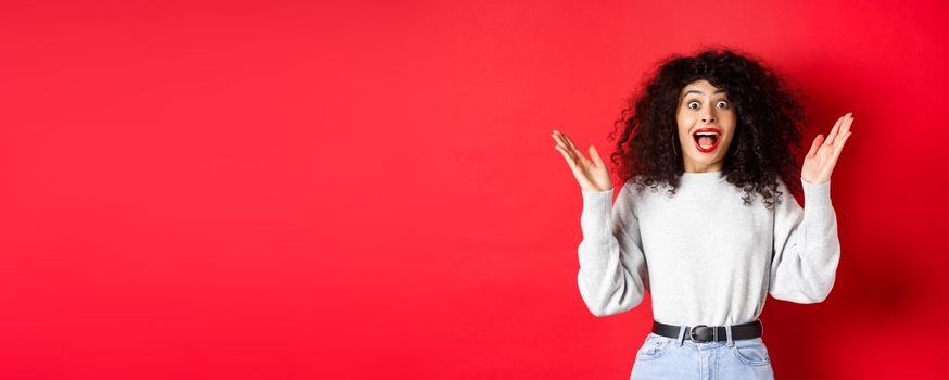 Image of happy and surprised curly woman in makeup and sweatshirt, raising hands up and rejoicing from good news, standing on red background.