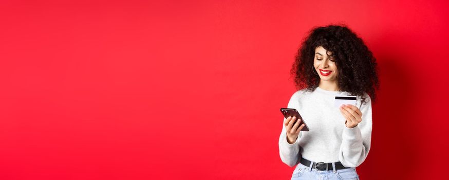 E-commerce and online shopping concept. Attractive caucasian woman paying for purchase in internet, holding smartphone and credit card, red background.