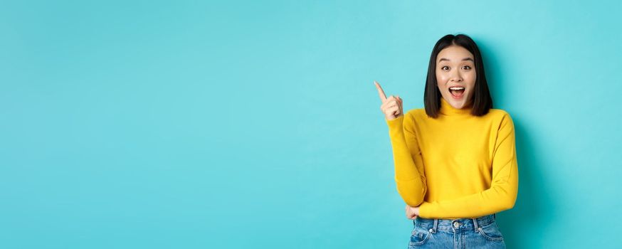 Shopping concept. Attractive korean woman smiling amazed, pointing finger left, showing good deal banner, standing against blue background.