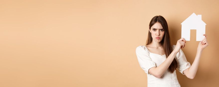 Real estate. Angry girl showing house cutout and frowning upset, complaining on apartment, standing against beige background.