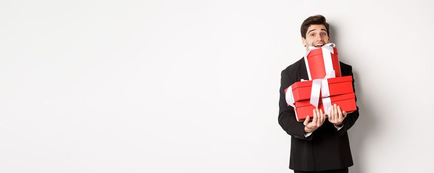 Concept of christmas holidays, celebration and lifestyle. Image of happy man in suit carry presents for new year, holding boxes with gifts and smiling, standing against white background.