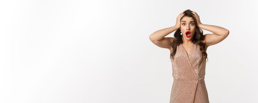 Celebration and party concept. Shocked and scared woman grab head and screaming, wearing glamour dress, standing over white background.