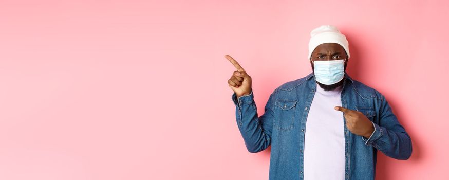 Coronavirus, lifestyle and global pandemic concept. Angry and disappointed african-american man in face mask pointing left, staring at camera displeased, pink background.