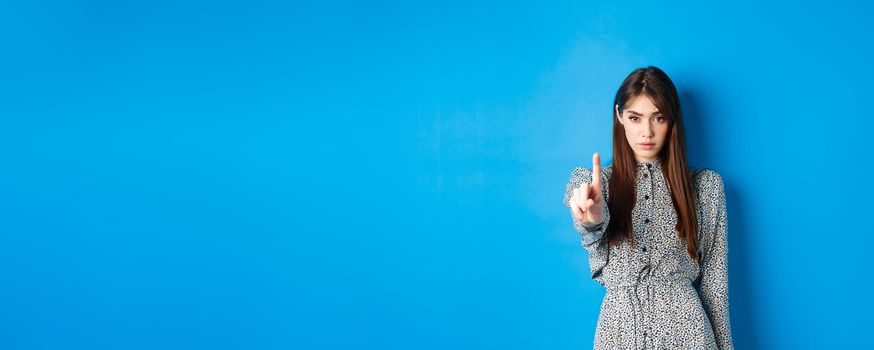 Serious woman looking confident and shaking finger, say no, stop you, disagree and disapprove, standing in dress on blue background.