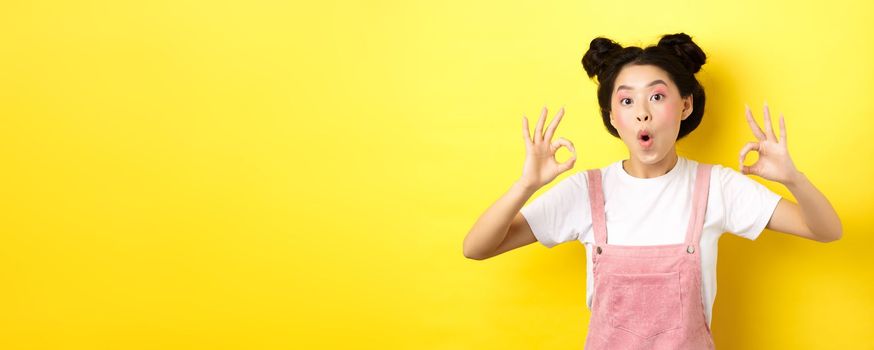 Excited beauty girl with bright makeup and summer clothes, showing okay signs and say wow, praise good promo deal, approve advertisement, yellow background.